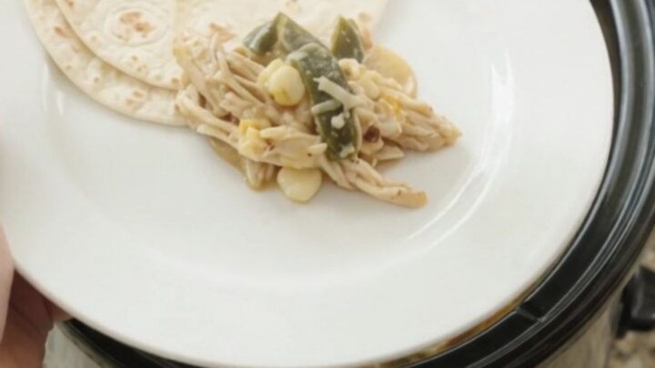 Creamy slow-cooker fiesta chicken with shredded chicken, hominy, and melted Monterey Jack in a black crockpot.