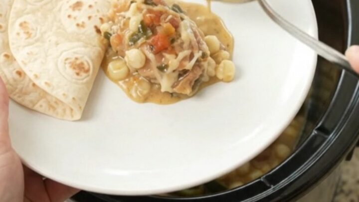 Creamy Crockpot chicken with poblanos and hominy in a black slow cooker, topped with melted cheese and salsa.