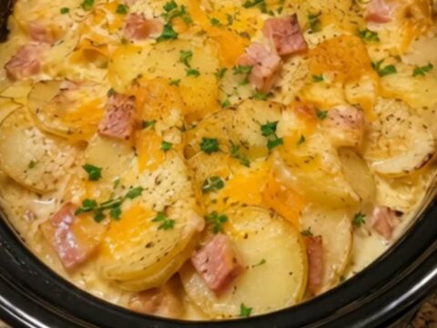 A close-up of creamy, cheesy scalloped potatoes and ham in a black slow cooker, garnished with fresh green parsley.