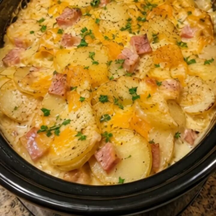 A close-up of creamy, cheesy scalloped potatoes and ham in a black slow cooker, garnished with fresh green parsley.