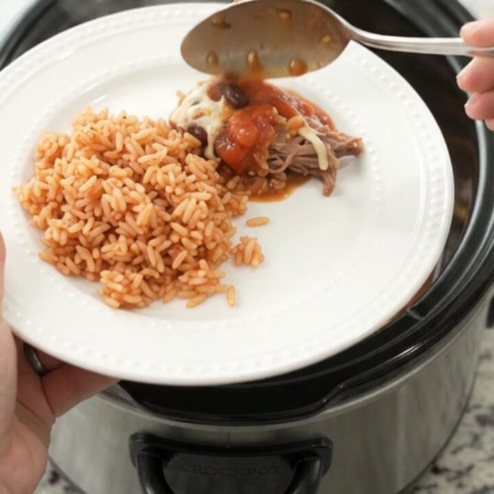 Slow cooker filled with shredded beef barbacoa, black beans, corn, and melted Oaxaca cheese, with steam rising.