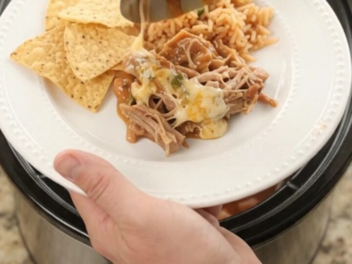 Slow cooker filled with shredded pork in red enchilada sauce and melted cheese, with a spoon lifting a bite.