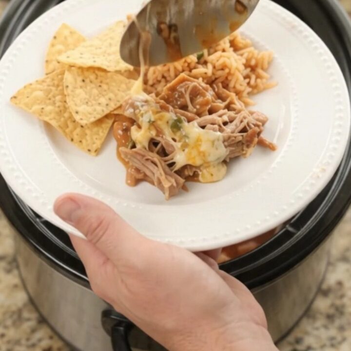 Slow cooker filled with shredded pork in red enchilada sauce and melted cheese, with a spoon lifting a bite.