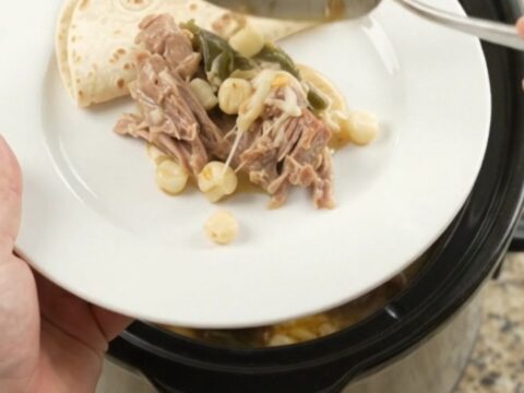 Creamy pork carnitas with hominy and poblano in a slow cooker, topped with melted cheese and steam rising.