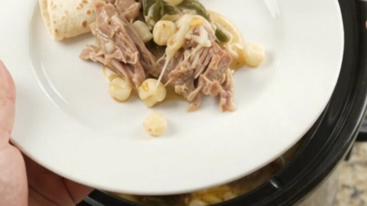 Creamy pork carnitas with hominy and poblano in a slow cooker, topped with melted cheese and steam rising.