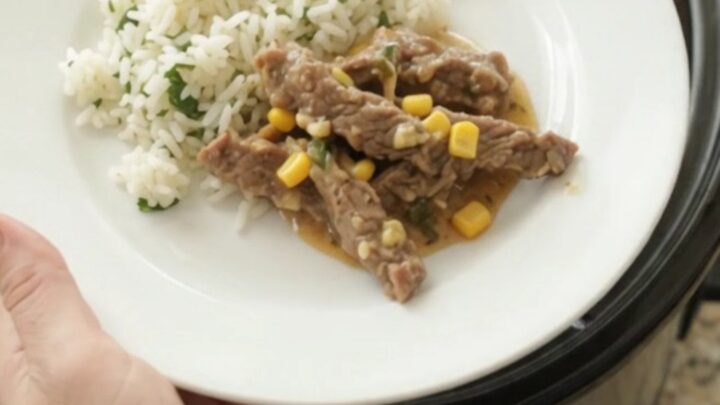 A slow cooker filled with tender skirt steak strips in a creamy cheddar and green chile sauce, with corn visible.