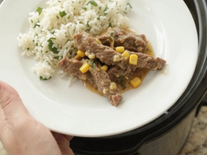 A slow cooker filled with tender skirt steak strips in a creamy cheddar and green chile sauce, with corn visible.