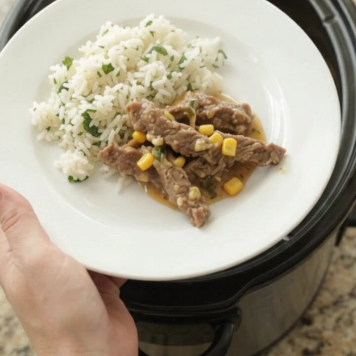 A slow cooker filled with tender skirt steak strips in a creamy cheddar and green chile sauce, with corn visible.