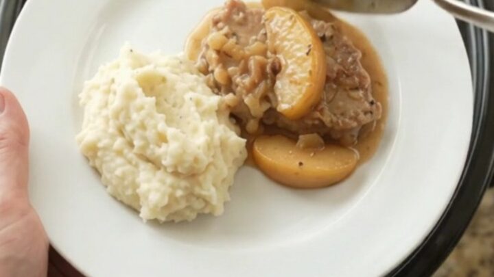 Slow cooker pork chops with apples in a creamy butter sauce, garnished with fresh thyme on a white plate.