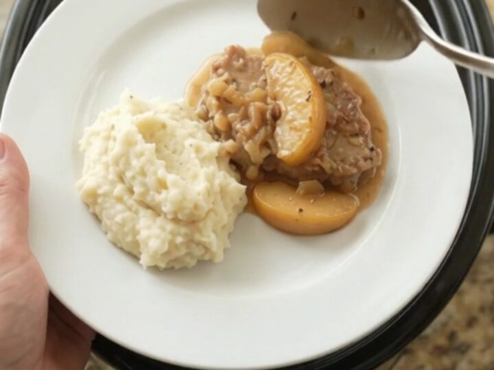 Slow cooker pork chops with apples in a creamy butter sauce, garnished with fresh thyme on a white plate.