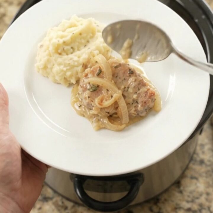 Crockpot full of tender pork chops in a creamy onion and Gruyère sauce, with steam rising.