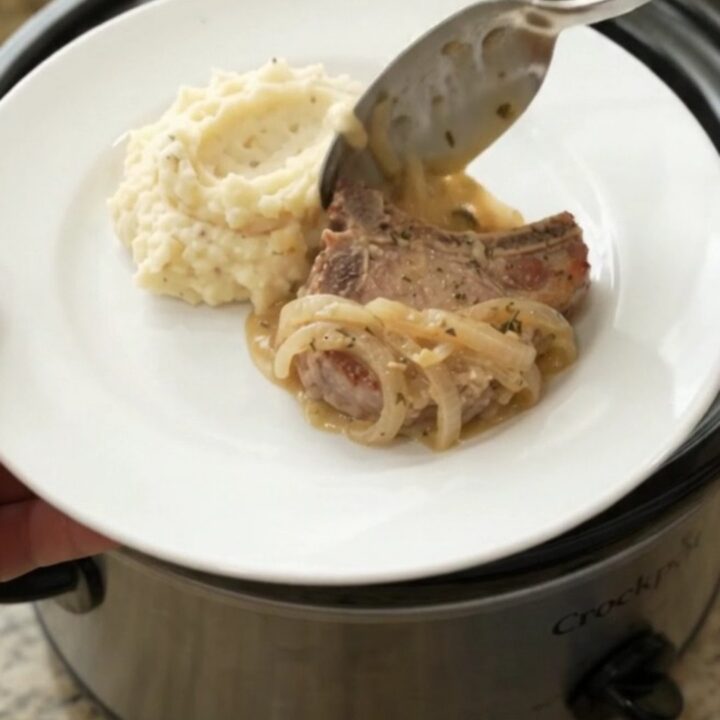 Slow cooker filled with creamy Gruyère onion sauce and tender pork chops, garnished with thyme.