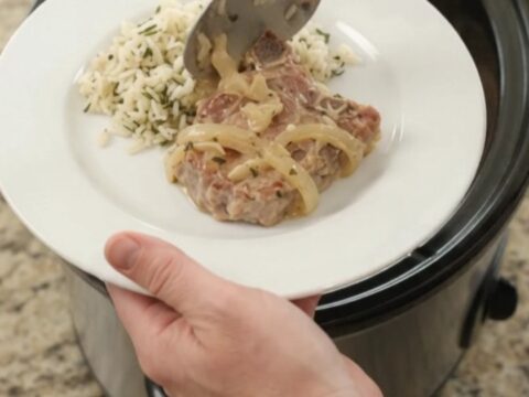 Slow-cooker Parmesan French onion ribeye pork chops in a black crockpot, topped with melted cheese and onions.