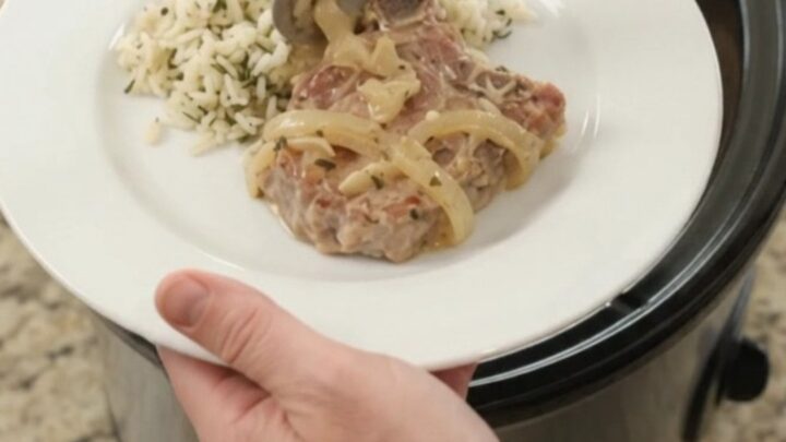 Slow-cooker Parmesan French onion ribeye pork chops in a black crockpot, topped with melted cheese and onions.