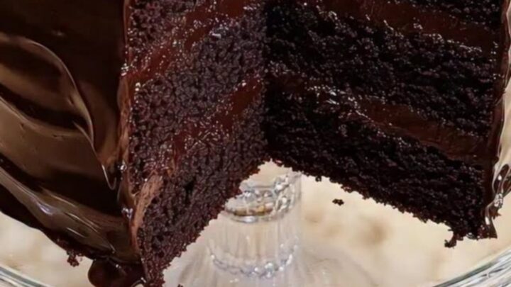 A triple-layer dark chocolate cake on a beaded glass stand with a slice removed to show the moist crumb.