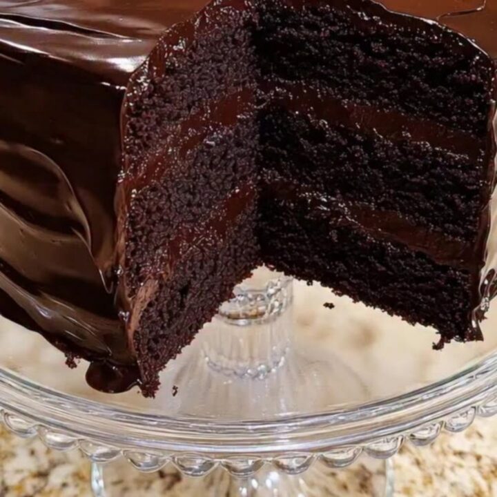 A triple-layer dark chocolate cake on a beaded glass stand with a slice removed to show the moist crumb.