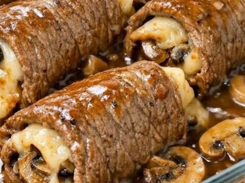 A glass baking dish filled with six golden-brown beef roll-ups smothered in a rich, dark mushroom gravy.