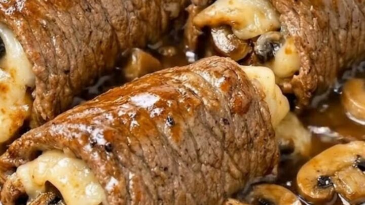 A glass baking dish filled with six golden-brown beef roll-ups smothered in a rich, dark mushroom gravy.