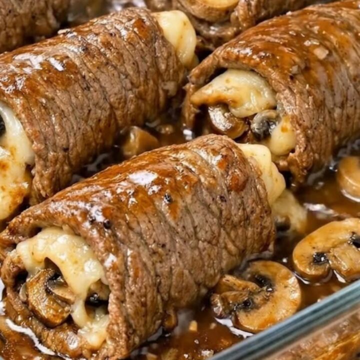A glass baking dish filled with six golden-brown beef roll-ups smothered in a rich, dark mushroom gravy.