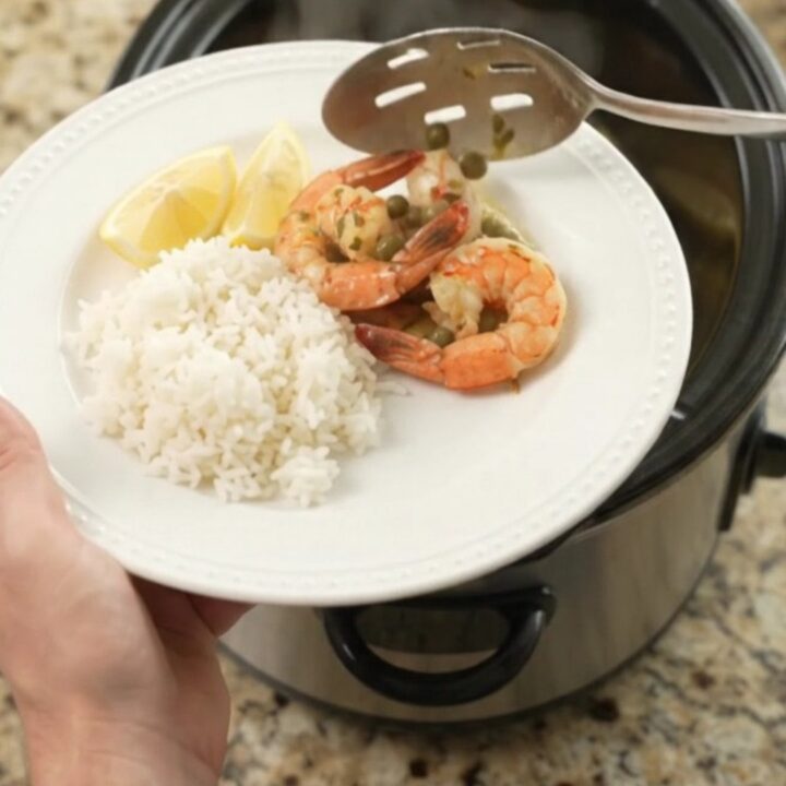 Slow cooker full of snow crab clusters and shrimp in a glossy chili-lime butter sauce with lemon slices and capers