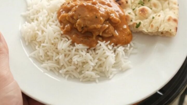 A slow cooker full of tender chicken thighs in a glossy orange-red makhani sauce with steam rising.