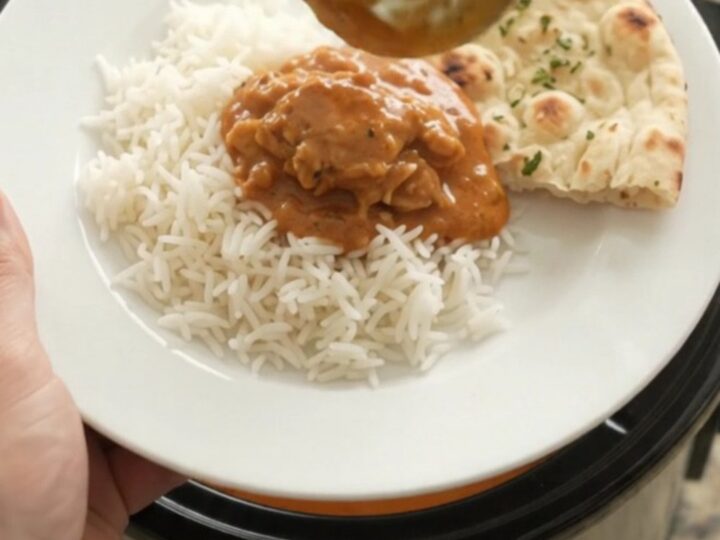 A slow cooker full of tender chicken thighs in a glossy orange-red makhani sauce with steam rising.