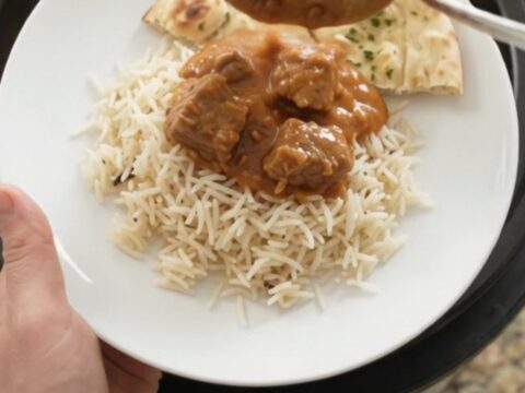 A slow cooker full of tender lamb cubes in a rich orange-red makhani sauce, with steam rising and a spoon lifting a bite.