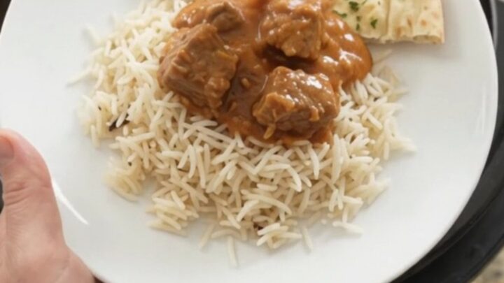 A slow cooker full of tender lamb cubes in a rich orange-red makhani sauce, with steam rising and a spoon lifting a bite.