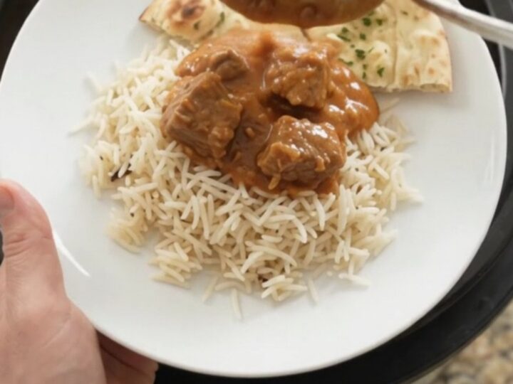 A slow cooker full of tender lamb cubes in a rich orange-red makhani sauce, with steam rising and a spoon lifting a bite.