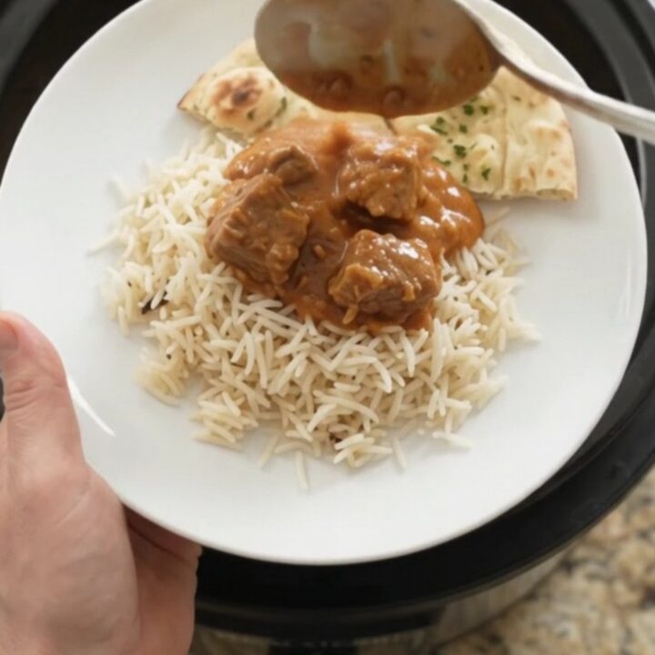 A slow cooker full of tender lamb cubes in a rich orange-red makhani sauce, with steam rising and a spoon lifting a bite.