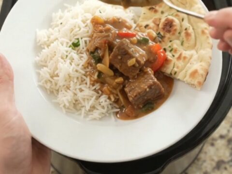 A slow cooker filled with red curry beef brisket, garnished with fresh cilantro and served with rice on the side.