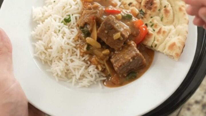 A slow cooker filled with red curry beef brisket, garnished with fresh cilantro and served with rice on the side.