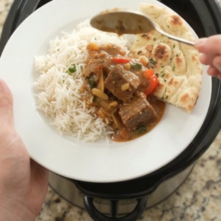 A slow cooker filled with red curry beef brisket, garnished with fresh cilantro and served with rice on the side.