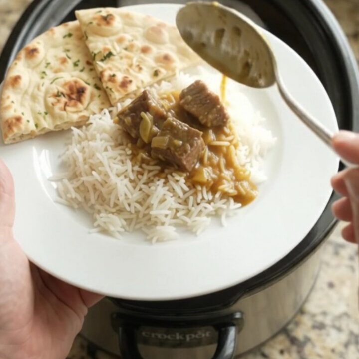 Slow cooker garlic butter korma beef brisket in a black ceramic pot, garnished with fresh herbs