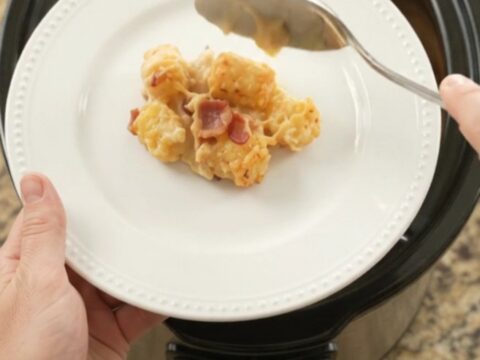 Golden bubbling cheddar tater tot bake in a black slow cooker, with a spoon lifting a cheesy scoop.