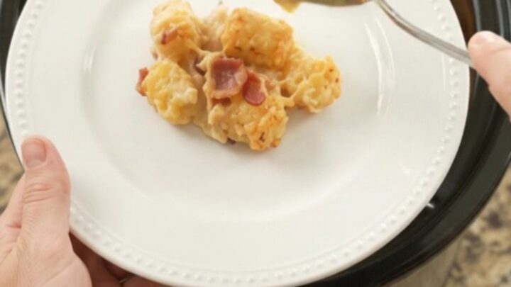 Golden bubbling cheddar tater tot bake in a black slow cooker, with a spoon lifting a cheesy scoop.
