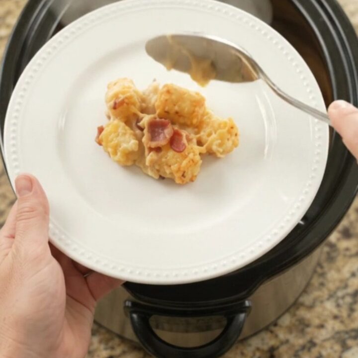 Golden bubbling cheddar tater tot bake in a black slow cooker, with a spoon lifting a cheesy scoop.