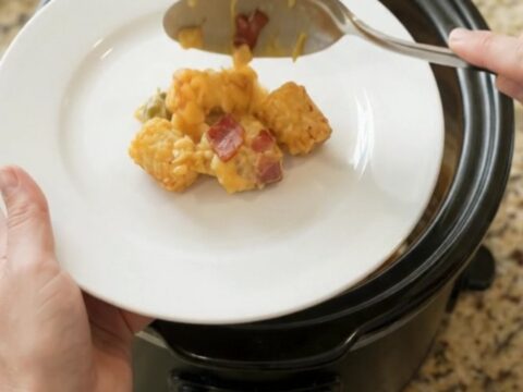 Bubbling golden cheddar green chile tater tot bake in a black Crockpot, with steam rising and a spoon lifting a cheesy scoop.