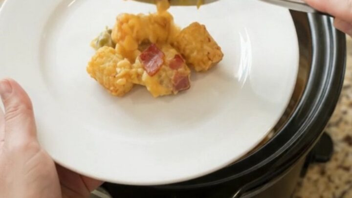 Bubbling golden cheddar green chile tater tot bake in a black Crockpot, with steam rising and a spoon lifting a cheesy scoop.