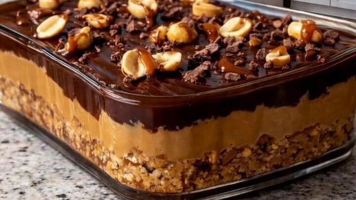 A clear glass dish filled with chocolate caramel peanut bars on a granite countertop, showing distinct layers.
