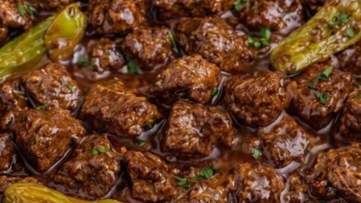 A close-up of tender beef cubes in a rich, dark gravy with whole pepperoncini peppers inside a slow cooker.