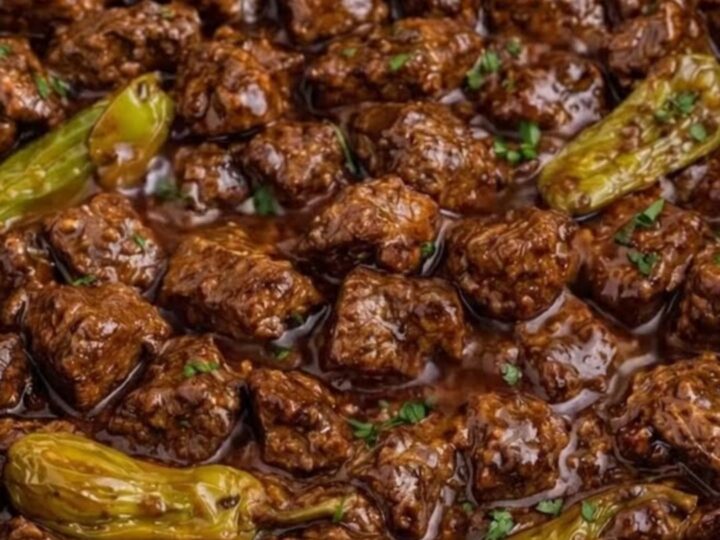 A close-up of tender beef cubes in a rich, dark gravy with whole pepperoncini peppers inside a slow cooker.