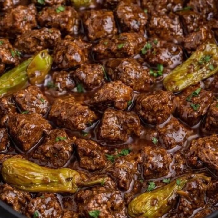 A close-up of tender beef cubes in a rich, dark gravy with whole pepperoncini peppers inside a slow cooker.