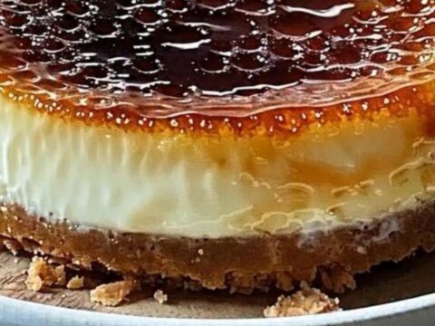 A close-up view of a silver spoon cracking the golden, brittle sugar crust of a creamy cheesecake.