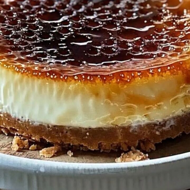 A close-up view of a silver spoon cracking the golden, brittle sugar crust of a creamy cheesecake.