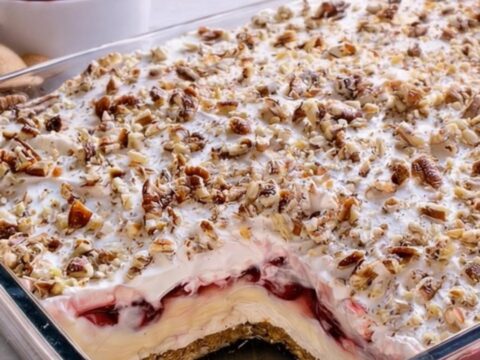 A glass dish filled with layers of graham crust, cream cheese, and cherry filling topped with pecans.