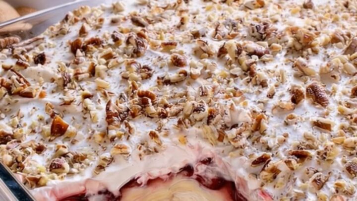 A glass dish filled with layers of graham crust, cream cheese, and cherry filling topped with pecans.