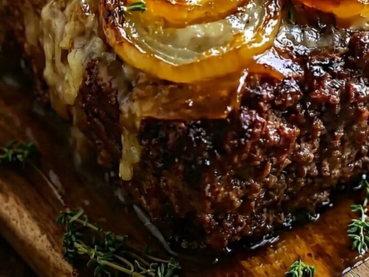 A close-up of a juicy beef meatloaf topped with golden caramelized onions and fresh thyme on a rustic wooden board.