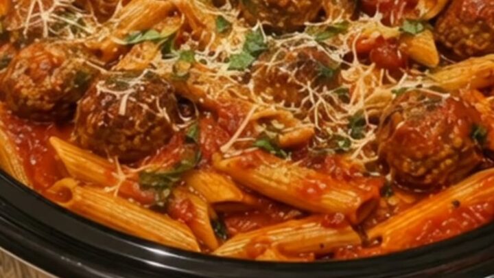 A steaming crockpot filled with penne pasta and meatballs in a rich red marinara sauce, garnished with fresh parsley.