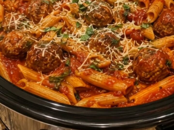 A steaming crockpot filled with penne pasta and meatballs in a rich red marinara sauce, garnished with fresh parsley.